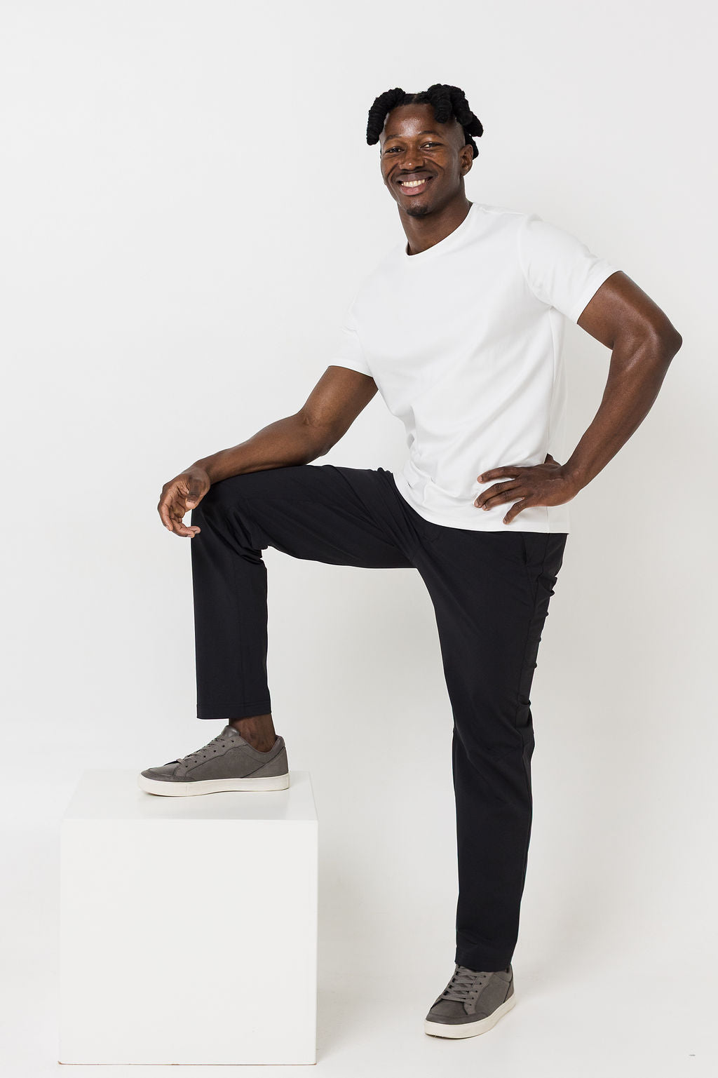 Man sitting on a white cube wearing a white t-shirt and black pants against a white background